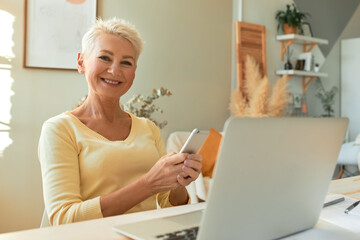Portrait of cheerful middle aged businesswoman in casual clothing working from home, sitting in front of open laptop, holding smart phone, making business calls, smiling broadly at camera