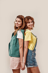 Portrait of two attractive young girls, twin sisters in casual wear smiling at camera, holding hands, posing together, standing back to back isolated over light background