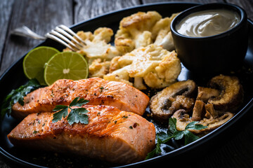 Roasted salmon steaks with fried cauliflower, fried brown champignons, lime and mayonnaise on wooden table
