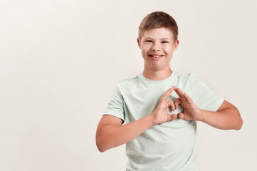 Joyful disabled boy with Down syndrome smiling at camera, making heart shape with his hands while standing isolated over white background