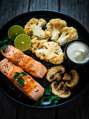 Roasted salmon steaks with fried cauliflower, fried brown champignons, lime and mayonnaise on wooden table

