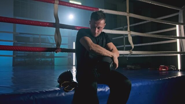 Young athlete getting ready for fight sitting on ring. Man wearing boxing gloves before training in slow motion. MMA fighter preparing. Smoky gym. Sport concept in 4K, UHD