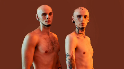 Portrait of young half naked twin brothers with tattoos and piercings looking at camera, posing together, standing isolated over orange background