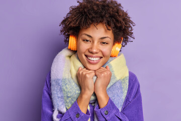 Close up portrait of lovely dark skinned millennial girl keeps hands under chin smiles broadly enjoys leisure time wears winter clothes listens pleasant music isolated over purple background