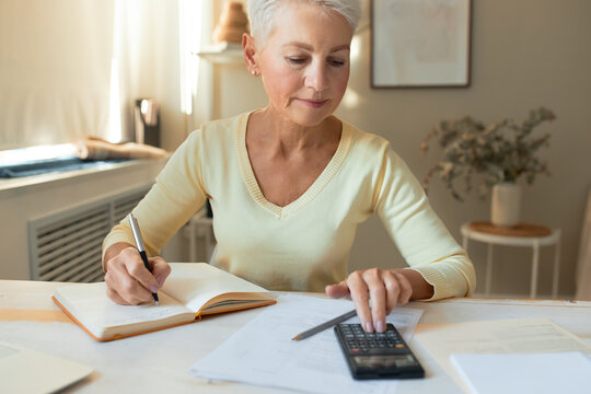 Indoor image of attractive serious retired female posing indoors with notebook and calculator on table, making notes while managing family budget, writing down numbers, calculating expenses