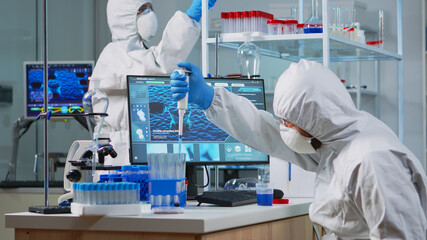 Research scientist in protection suit using micropipette filling test tubes in lab. Team of...