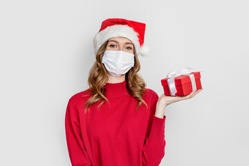 New Year's gifts 2021 during the coronavirus quarantine pandemic. Girl student in a red sweater put on in a medical mask and santa hat holds a gift box isolated on white studio background.