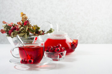 Morning autumn teatime with red herbal tea in transparent tea pot, cup with hawthorn, rosehip berry, dried leaves in sun beam in soft light white interior.