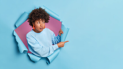 Surprised dark skinned Afro American woman looks with great wonder and advertises item shows copy space for your promotion dressed in long sleeved jumper poses through paper hole. Breakthrough - obrazy, fototapety, plakaty