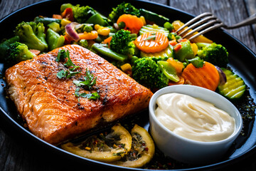 Fried salmon fillet with mix of vegetables, lemon and mayonnaise served on black plate on wooden...