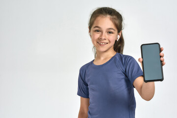 Fototapeta premium Portrait of little sportive girl child wearing earphones, smiling at camera and showing smartphone with blank screen, standing isolated over white background