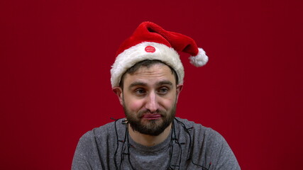 The man is sitting and a Christmas hat is dancing on his head. Santa's bearded helper on red background