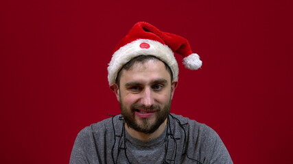 The man is sitting and a Christmas hat is dancing on his head. Santa's bearded helper on red background
