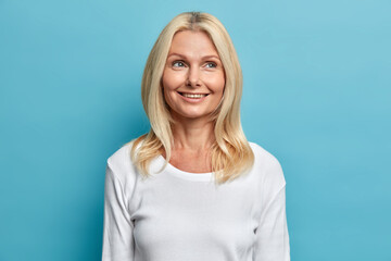 Portrait of beautiful dreamy blonde middle aged woman with pleased face expression feels good recalls nice memories concentrated above wears casual white jumper poses against blue background.