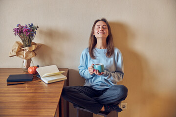 Young cheerful confident and happy woman is enjoying tea while reading book at her kitchen at home