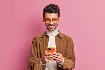 Studio shot of European man blogger types text messages on smartphone smiles pleasantly wears...