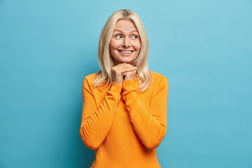 Image of mature blonde gorgeous woman looks gladfully aside keeps hands under chin smiles gently admires something wears orange jumper isolated over blue background. Age emotions beauty concept