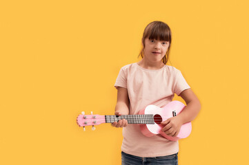 Cheerful disabled girl with Down syndrome looking at camera while playing ukulele, standing...