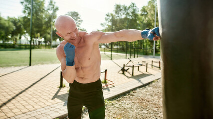 Focused mature man boxing with black punching bag, having workout at street gym yard