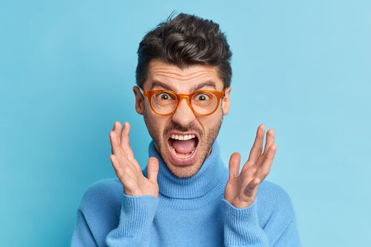 Close up portrait of displeased young man raises hands and screams loudly being annoyed with something keeps mouth widely opened wears optical glasses and turtleneck poses indoor. People irritation