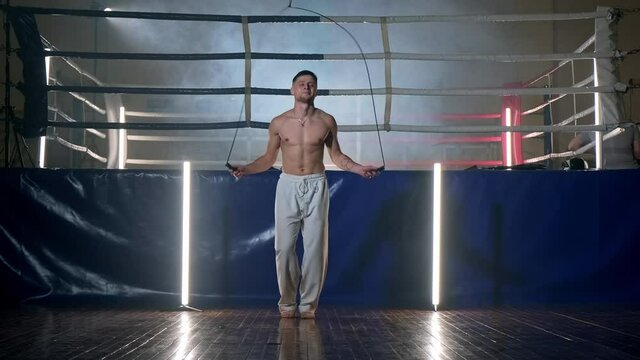 Fighter doing jump rope exercises in dark gym with light stands at background. Shirtless male fighter with skipping rope. Warming up and training of taekwondo fighter. Full length in 4K, UHD