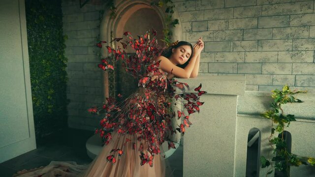 Portrait of beautiful fantasy woman fairy tale queen bride. Princess Girl fashion model in old room castle. Long trendy creative dress decorated with artificial red butterflies. Elegant lady dreaming