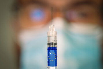 Close-up of hypodermic syringe with the European Union flag and a blurred doctor on the background. Selective focus. Concept of Covid vaccination campaign in Europe