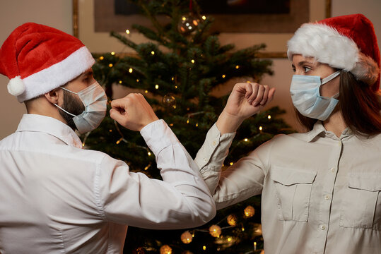 Elbow Greeting To Avoid The Spread Of Coronavirus (COVID-19) Against A Background Of A Christmas Tree At Home. Man And A Woman In Medical Masks And Santa Hats Instead Of A Hug Or Handshake Bump Elbows