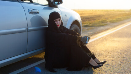 Woman driver sits on the asphalt near a broken car.
