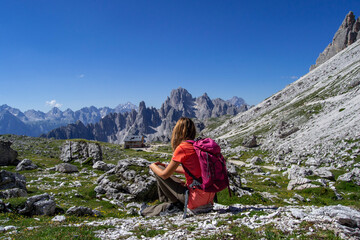 Naklejka na ściany i meble Young woman with backpack enjoying breathtaking view of Dolimites Alps mountain landscape sitting on a rock. Travel concept back view