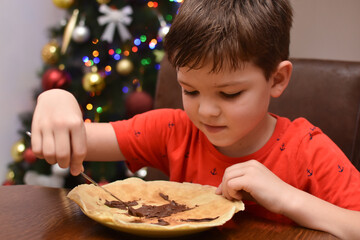 Little boy eating pancakes in front of traditional Christmas tree. Child enjoy eating pancake in Christmas Eve holiday celebration
