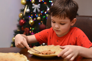 Little boy eating pancakes in front of traditional Christmas tree. Child enjoy eating pancake in Christmas Eve holiday celebration