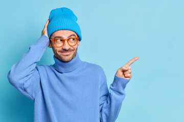 Studio shot of glad male teenager in fashionable clothes looks with interest and points away on...