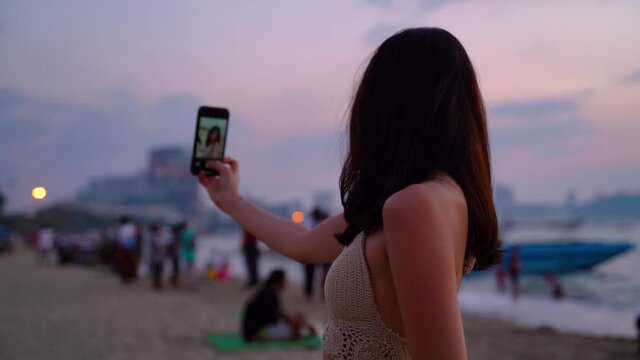 Young woman taking a selfie photo in sunset tropical beach sea view background with her mobile phone. Beautiful Asian girl photographing herself with smartphone - 4K Quality Footage