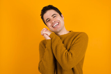 Dreamy Young handsome Caucasian man wearing yellow sweater against orange wall with pleasant expression, closes eyes, keeps hands crossed near face, thinks about something pleasant