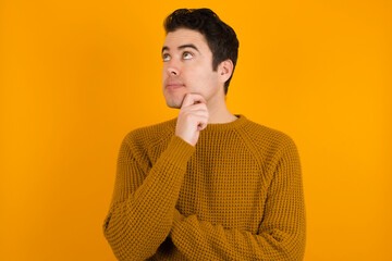 Portrait of thoughtful Young handsome Caucasian man wearing yellow sweater keeps hand under chin, looks away trying to remember something or listens something with interest. Youth concept.