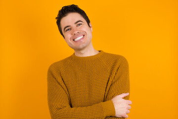 People, lifestyle, youth and happiness concept. Shy pretty Young handsome Caucasian man wearing yellow sweater against orange wall, feeling happy hugging herself.