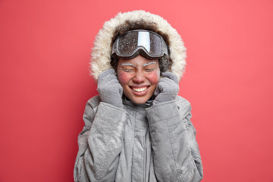 Photo of overjoyed woman wears hood of grey jacket smiles pleasantly has red face covered with frost goes skiing during December isolated over pink background. Frozen firl being not accustomed to cold