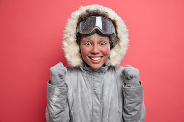 Emotive positive woman raises fists and cannot wait until going snowboarding expresses happy emotions enjoys winter time and active rest wears warm jacket and ski goggles. Cheerful female skier