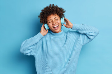 Carefree joyful dark skinned young woman listens audio track in headphones closes eyes and smiles broadly expresses happy emotions enjoys music isolated over blue background. Monochrome shot