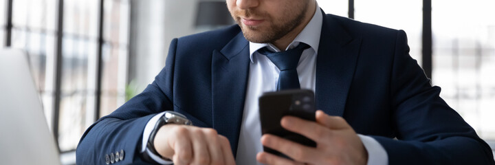 Horizontal banner confident businessman looking at wrist watch, holding smartphone, checking time, waiting for meeting, sitting at desk in office, planning workday, task management concept