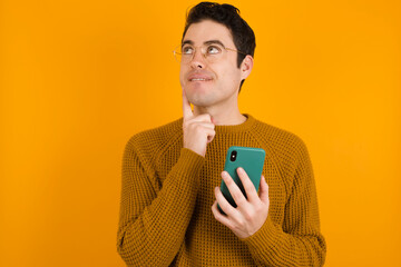 Image of a thinking dreaming Young handsome Caucasian man wearing yellow sweater against orange wall using mobile phone and holding hand on face. Taking decisions and social media concept.