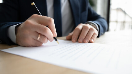 Close up business owner business man wearing suit signing documents, filling form, making successful deal, sitting at office desk, employee candidate signing job agreement after interview