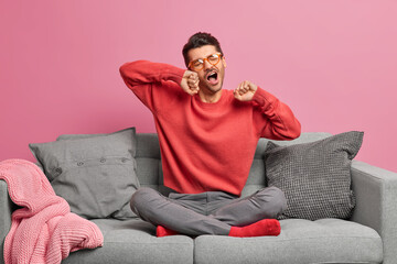 Indoor shot of sleepy European man stretches and sits in lotus pose on comfortable sofa wants to sleep enjoys cozy domestic atmosphere isolated over pink background. Staying at home concept.