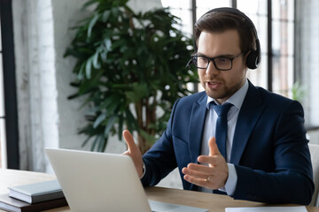 Close up focused employee wearing glasses and headset consulting client, using laptop, confident businessman looking at laptop screen, making video call to business partner, chatting online