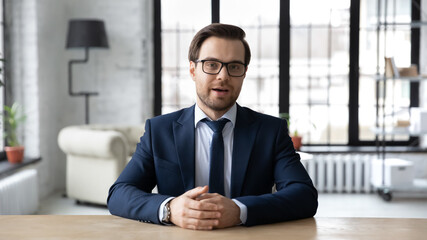 Head shot confident businessman wearing suit and glasses speaking looking at camera, sitting at...