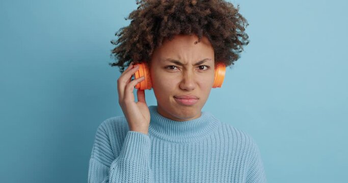 Puzzled displeased dark skinned young woman listens song shows dislike gesture keeps thumb down doesnt like music from playlist wears wireless stereo headphones poses against blue background