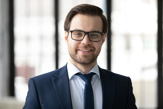 Head shot portrait close up smiling confident businessman wearing glasses and suit, profile picture happy successful company owner executive employee looking at camera, standing posing in office