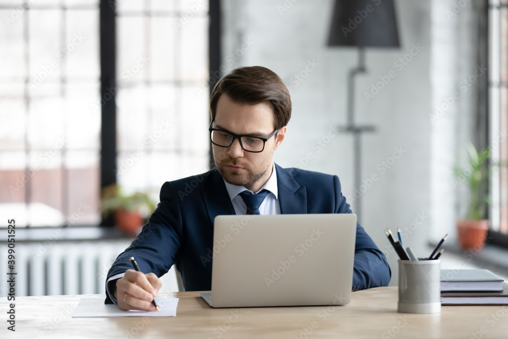 Poster Confident businessman wearing suit and glasses using laptop, writing notes or financial report, sitting at desk in office, focused young entrepreneur working with documents or marketing plan