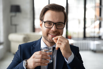 Head shot portrait smiling businessman wearing glasses taking meds, medication pills in office, holding glass of water, looking at camera, happy man employee drinking daily vitamins, supplements